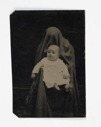 Portrait of baby in white (seated on lap of shrouded figure)