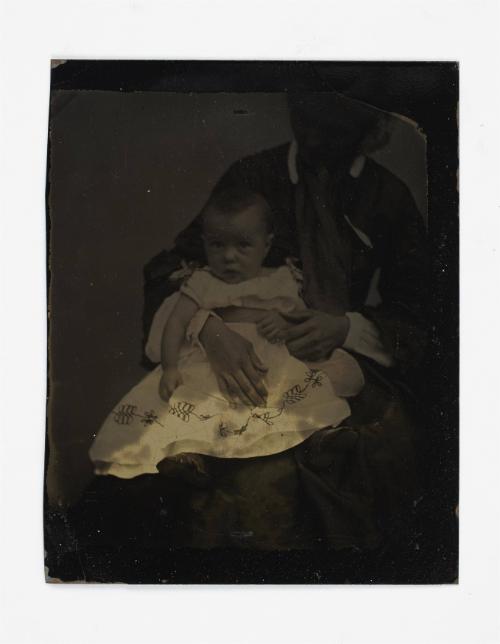 Portrait of baby  in white embroidered dress (seated on lap supported by defaced figure)