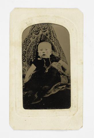 Portrait of baby in black dress (seated on lap of shrouded figure)