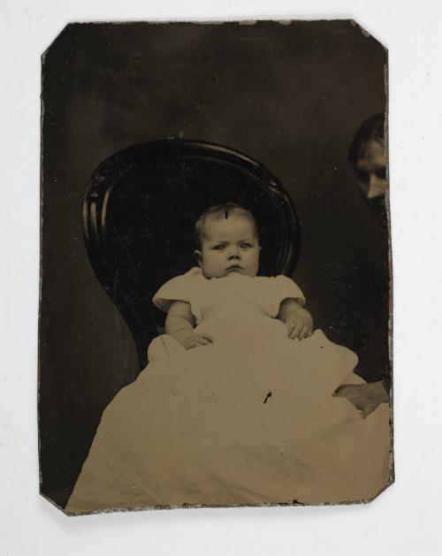 Portrait of baby in long white dress (seated on Queen Anne chair supported, by cropped woman)