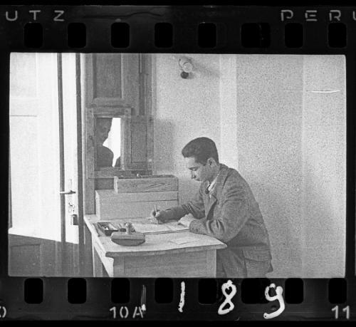 Young man working at a desk (with man looking through the open window)