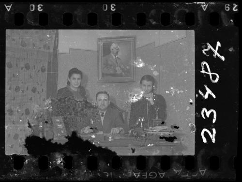 Man sitting at desk with two women, beneath a portrait of Mordechai Chaim Rumkowksi