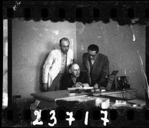 Three men (probably members of the Judenrat) reviewing documents at a desk. Man in the white coat wears a Jewish policeman's armband.
