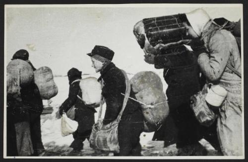 Boy in cap, with satchel and sack tied to back, walking with winter deportation crowd -side-profile