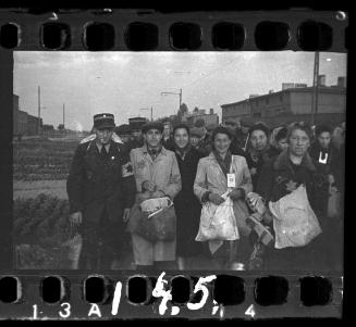 Policemen and residents carrying belongings in crowd assembled for deportation