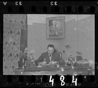 Man working at a desk beneath a portrait of Chaim Rumkowski, the Elder of the Jewish Council