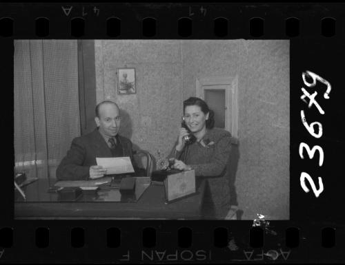 Man with paper and woman with phone sitting at a desk in an office, with a portrait of Mordechai Chaim Rumkowksi on the wall