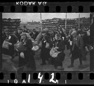 Deportation group with belongings passing piles of wood