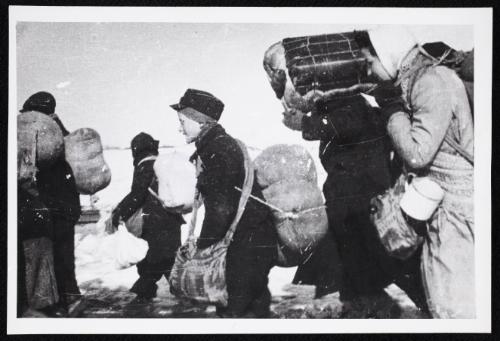 Boy in cap, with satchel and sack tied to back, walking with winter deportation crowd -side-profile