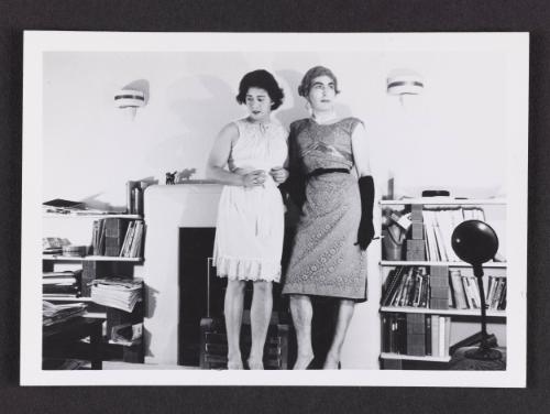 [Irène and friend standing by fireplace, location unknown]
