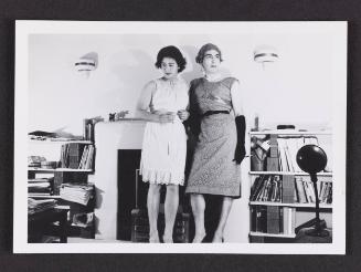 [Irène and friend standing by fireplace, location unknown]
