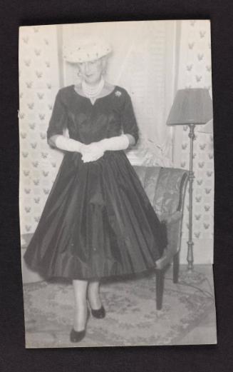 [In hat and pearls standing in front of armchair, location unknown]
