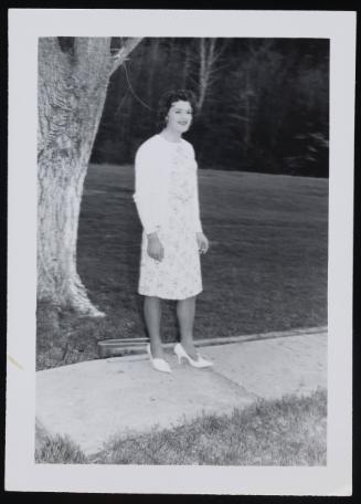 [Brunette standing on the sidewalk in a white dress and sweater, location unknown]
